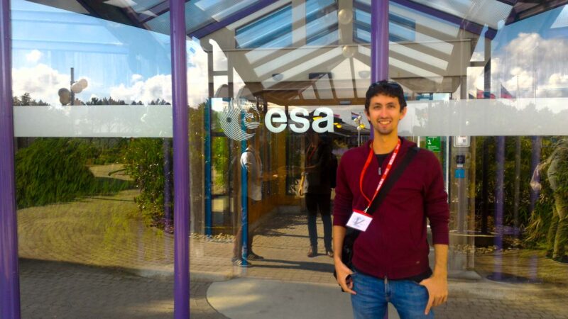 Stefano in front of a building with glass doors, which show the logo of ESA