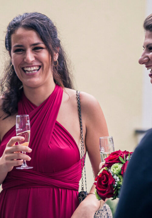 Isi in a red neckholder dress with a glass of champagne laughing with Luise and other people around